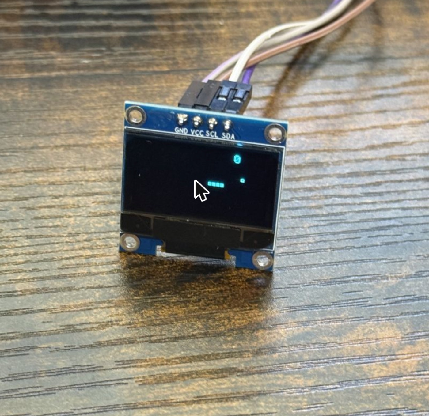 A small blue OLED breakout board wired to jumper leads on a wooden desk, showing a few pixels of a Snake game in progress.